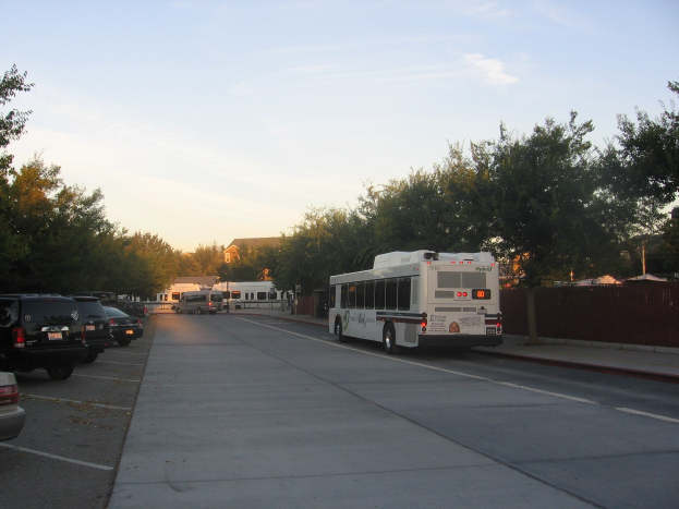 Shuttle-Bus fährt auf einer Straße mit parkenden Autos, Bäumen, Gebäuden und einem klaren blauen Himmel im Hintergrund.