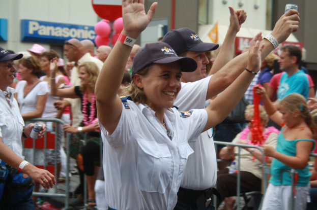 Eine Gruppe von Menschen, einige mit Mützen und Dosen, steht und sitzt vor einer Menge bei einem Umzug, mit Geländern, Luftballons und einem Gebäude mit Namensschildern im Hintergrund.