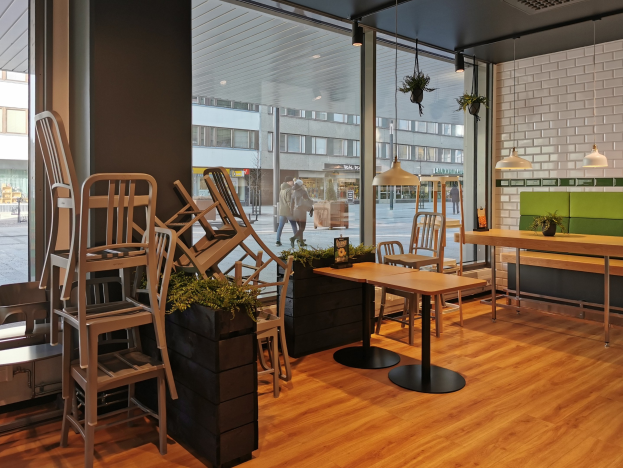 Ein Restaurant mit Holztischen und -stühlen, das durch ein großes Fenster zu sehen ist, das in Berlin, Deutschland, mit Passanten, Gebäuden und Grünflächen draußen liegt.
