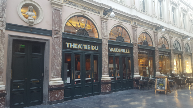 Theater du Vaudeville-Gebäude in Paris mit Türen, Fenstern, beleuchteter Beschilderung, Außenbestuhlung und einer Wandstatue.