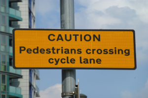 Gelbes Warnschild an einem Pfahl vor einem Gebäude, mit einem klaren blauen Himmel im Hintergrund, das 'Fußgänger überqueren Fahrradweg' anzeigt.