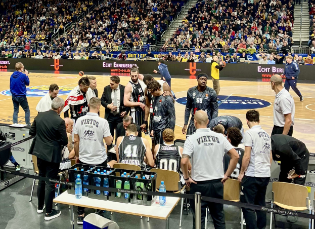 Eine Gruppe von Männern auf einem Basketballfeld während eines Spiels zwischen Virtus Berlin und Echt Berlin, umgeben von Zuschauern auf Stühlen und Stehplätzen, einem Fotografen mit Ausrüstung links daneben und Infotafeln im Hintergrund.