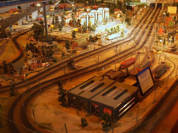 Eine detaillierte Miniatur-Modellbahnanlage in einem Museum, die Gebäude, Bäume, Eisenbahnschienen, Fahrzeuge, Masten, Lichter und andere Gegenstände enthält, die eine belebte Stadtlandschaft nachahmen.
