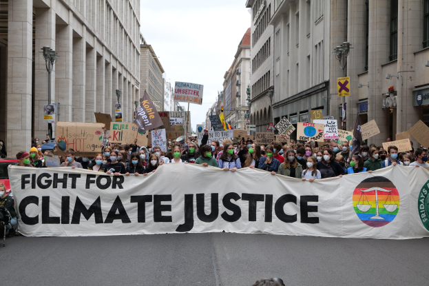 Eine große Gruppe von Menschen marschiert die Straße entlang und hält ein "Kämpfen für Klimagerechtigkeit"-Schild und verschiedene Plakate, trägt Masken, mit Gebäuden auf beiden Seiten der Straße und einem klaren blauen Himmel im Hintergrund.