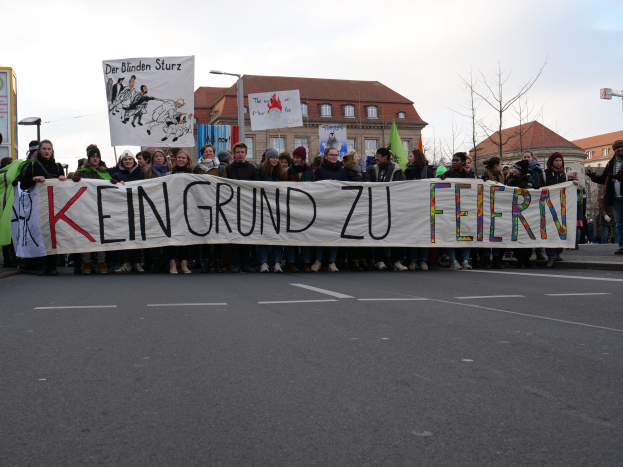Demonstranten mit einem Banner "Kein Grund zu Feiern" gegen deutsche Sparmaßnahmen auf einer Stadtstraße mit Gebäuden, Bäumen und einem klaren Himmel.
