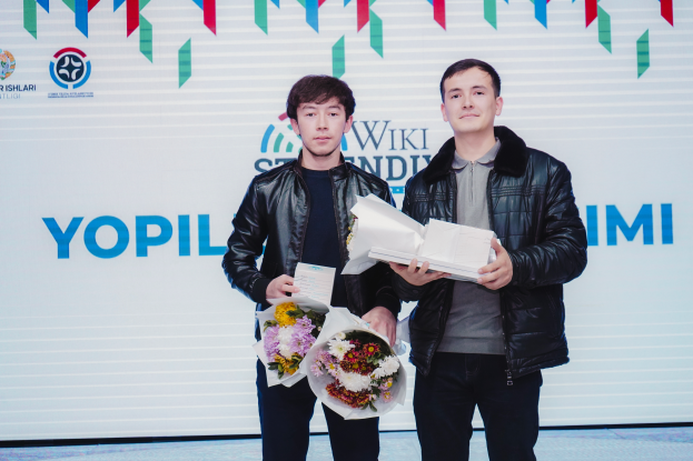 Zwei Männer in schwarzen Jacken stehen nebeneinander auf einer Bühne, jeder hält einen Blumenstrauß in der Hand, mit einem weißen Banner im Hintergrund, auf dem "Yopil International Film Festival" steht.