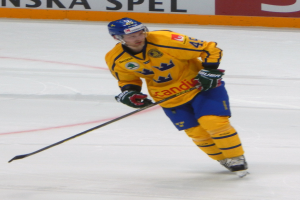 Eishockeyspieler in Bewegung auf dem Eis, trägt Helm, Handschuhe und Schlittschuhe, hält einen Hockeystock und hat eine Tafel mit Text im Hintergrund.