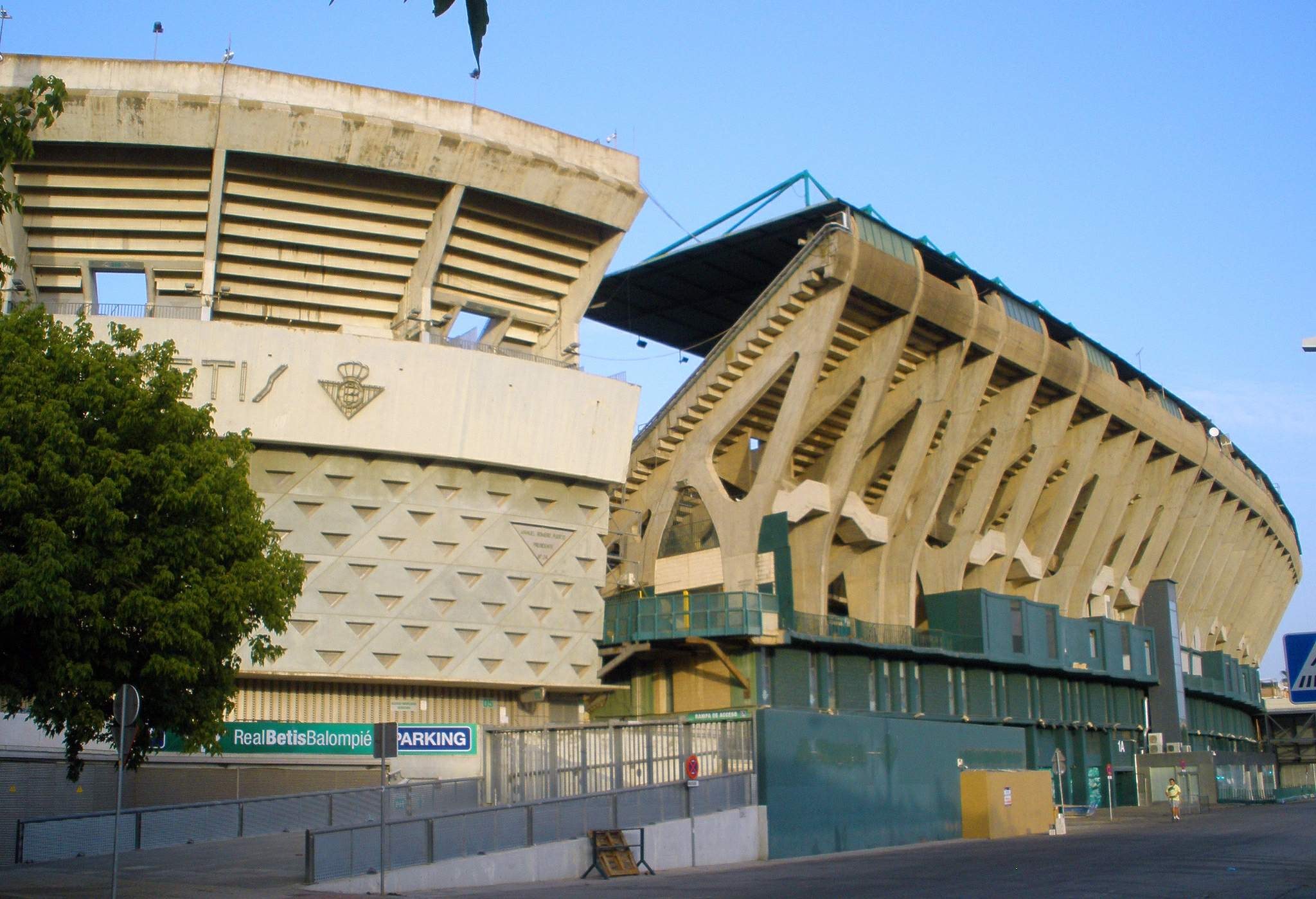 Großes Stadion mit zahlreichen parkenden Autos davor, umgeben von Gebäuden, Bäumen, Pfählen, Schildern, Gittern und einer Straße unter einem sichtbaren Himmel.