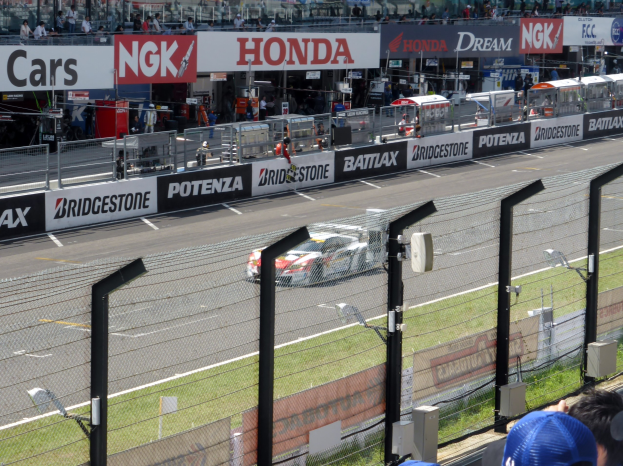Ein Rennwagen fährt auf einer Rennstrecke mit Zuschauern in den Tribünen, umgeben von Absperrungen und Lichtmasten.