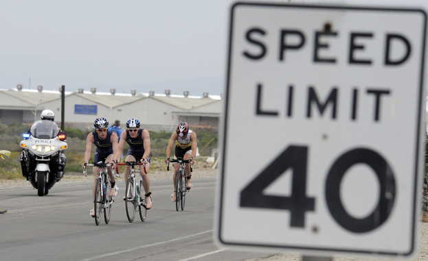 Gruppe von Radfahrern mit Helmen auf der Straße mit einem Geschwindigkeitsbegrenzungsschild, Gebäude und Bäume im Hintergrund unter einem klaren blauen Himmel.