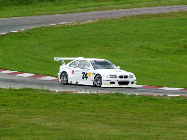 Ein weißer BMW M3 GT3 Rennauto, das auf einer Rennstrecke fährt, mit grünem Gras und Pflanzen im Hintergrund.
