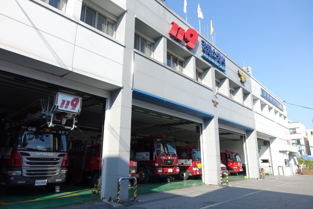 Feuerwache mit mehreren Feuerwehrautos vor der Tür, mit Fenstern, Säulen, Namensschildern und Flaggen im Vordergrund auf Stangen, vor anderen Gebäuden unter einem klaren blauen Himmel.