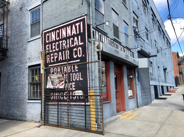 Außenansicht des Cincinnati Electrical Repair Co. Gebäudes mit einer Tafel, einem Metallzaun, einem Müllcontainer, Pfählen, Drähten, Bäumen und einem bewölkten Himmel.