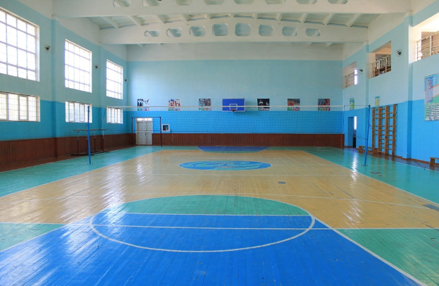 Großer Sportraum mit blau-weißem Boden, Basketballfeld, Netz, Stangen, Tür, Fenstern, Wandplakaten, Geländer und Dach.