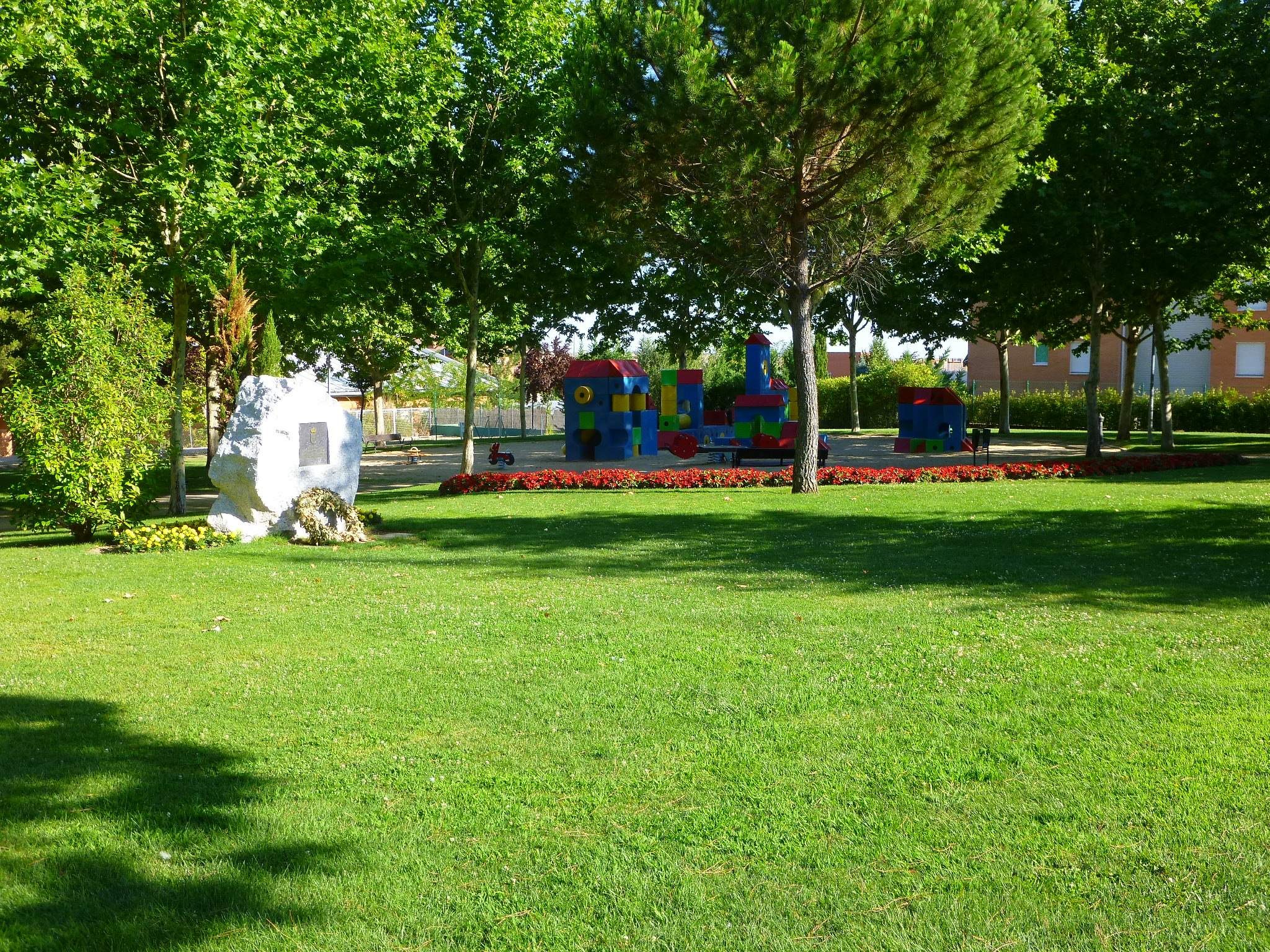 Ein Park mit grünem Gras, Bäumen, Pflanzen, Blumen, Steinen und bunten Spielgeräten, umgeben von Gebäuden unter einem klaren blauen Himmel.