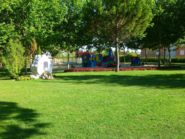 Ein Park mit grünem Gras, Bäumen, Pflanzen, Blumen, Steinen und bunten Spielgeräten, umgeben von Gebäuden unter einem klaren blauen Himmel.