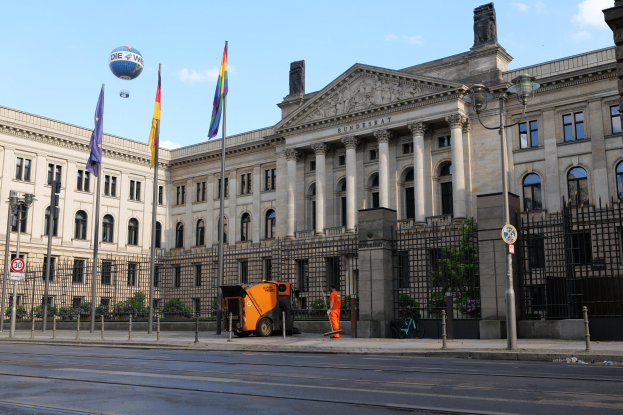 Großes modernes Gebäude mit vielen Fenstern, als Bundestag in Berlin, Deutschland identifiziert, umgeben von Straßeninfrastruktur und Füßgängern, mit Flaggen und Bäumen im Vordergrund und einem bewölktem Himmel im Hintergrund.