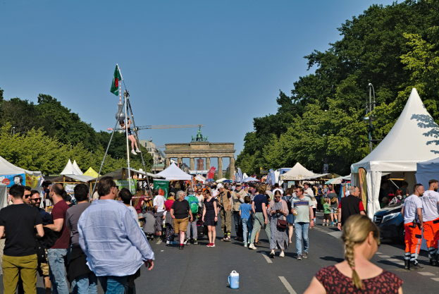 Eine Menschenmenge, die eine Straße mit Zelten, Fahrzeugen und Bäumen entlanggeht, mit einem Bogen und einem klaren blauen Himmel im Hintergrund und Polen mit Fahnen auf der linken Seite, wahrscheinlich das Oktoberfest in München, Deutschland darstellend.