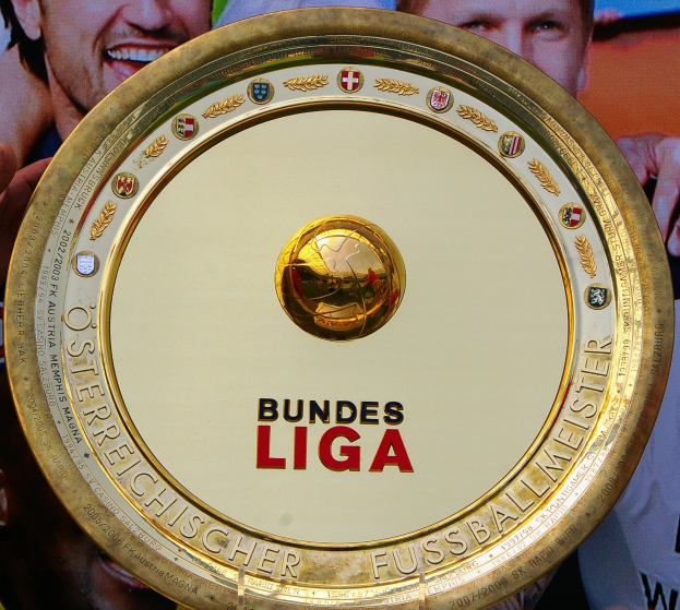 Ein Mann hält einen goldenen Schild mit der Aufschrift 'Bundesliga' hoch, im Hintergrund ein Banner mit drei Personen.