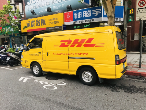 Ein gelber DHL-Lieferwagen parkt auf einer Straße neben Gebäuden, Bäumen, Schildern, Masten, Ampeln und einem Fussgänger.