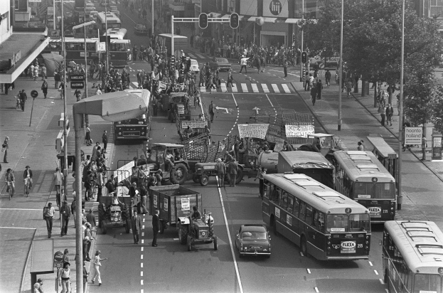 Schwarzes und weißes Foto einer belebten Stadtstraße mit Bussen, Autos, Lastwagen, Fußgängern, Gehwegen, Pfählen, Verkehrsampeln, Schildern, Bäumen und Gebäuden.