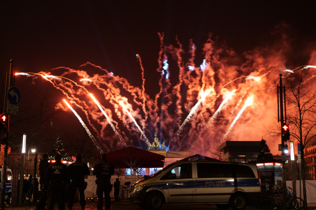 Polizeiauto vor Menschenmenge bei Silvesterfeier in Berlin geparkt, mit farbenfrohen Feuerwerken am Himmel.