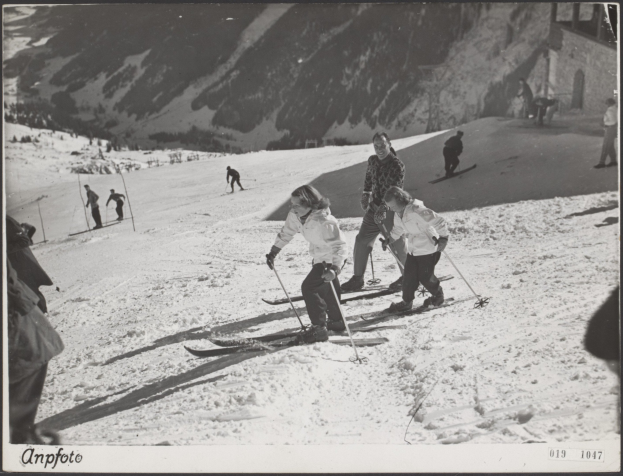 Gruppe von Menschen, die eine schneebedeckte Piste hinunterskien, Ski-Stöcke haltend, mit Hügeln und einem Gebäude im Hintergrund und Text unten.