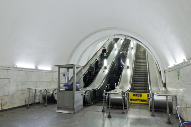 Rolltreppe in einer U-Bahn-Station mit Menschen, Geländern, Informationsschildern, Wandlampen und einem Einkaufswagen unten.