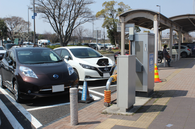 Elektroauto-Ladestation in Japan mit Autos, Verkehrskegeln, einer Person auf dem Gehweg, einem Schuppen, Polen, Lampen, Schildern, Bäumen, Pflanzen und einem sichtbaren Himmel.
