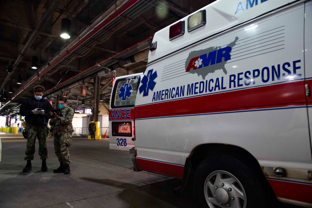 Ein Krankenwagen der American Medical Response (AMR) steht in einem Gebäude mit zwei maskierten Personen davor, einem Fahrzeug auf der linken Seite und mehreren Personen, Lichtern und Rohren im Hintergrund.