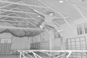 Ein Mann in der Luft, Arme ausgestreckt und Knie gebeugt, springt auf einem deckenbefestigten Trampolin in einer Turnhalle, dargestellt in Schwarz-Weiß.