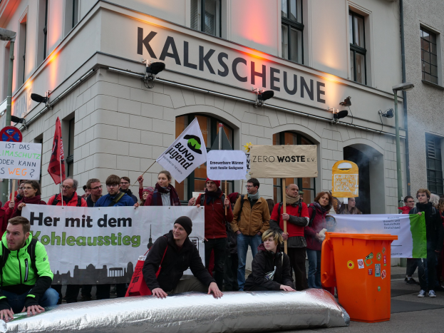 Eine Gruppe von Menschen protestiert vor einem Gebäude in Deutschland, mit Schildern und Plakaten, während zwei Personen im Vordergrund sitzen und ein Müllcontainer rechts zu sehen ist.