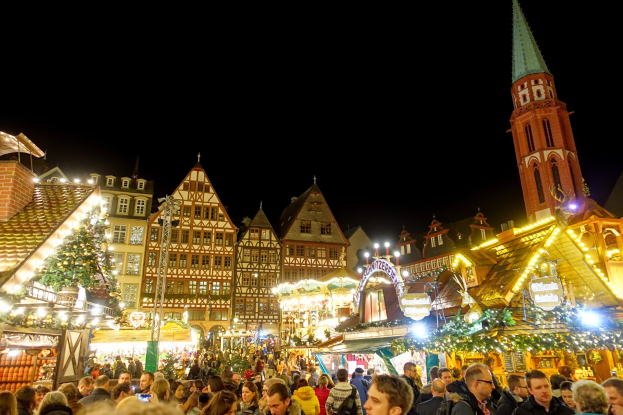 Ein geschäftiger Weihnachtsmarkt in Nürnberg, Deutschland mit Menschen um geschmückte Stände, festlicher Beleuchtung, Gebäuden mit Fenstern, einem Uhrturm und einem dunklen Himmel.