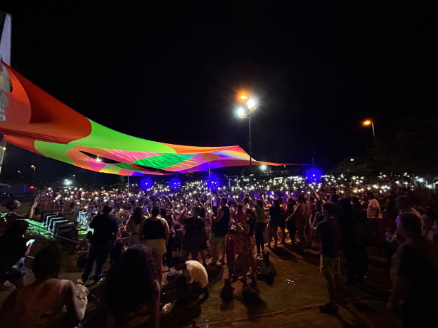 Eine große Menschenmenge steht vor einem hell erleuchteten Zelt in der Nacht, umgeben von farbigen Lichtern und beleuchteten Pfählen, mit einem Gebäude auf der linken Seite und dem Nachthimmel im Hintergrund.