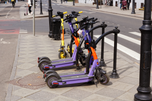 Eine Reihe von Elektro-Scootern, die auf der Straäenseite geparkt sind, mit Menschen, die auf dem Gehweg laufen und Fahrräder fahren, ein M"ulltonne im Vordergrund und Gebäude, Bäume, Pflanzen und andere Gegenstände im Hintergrund.