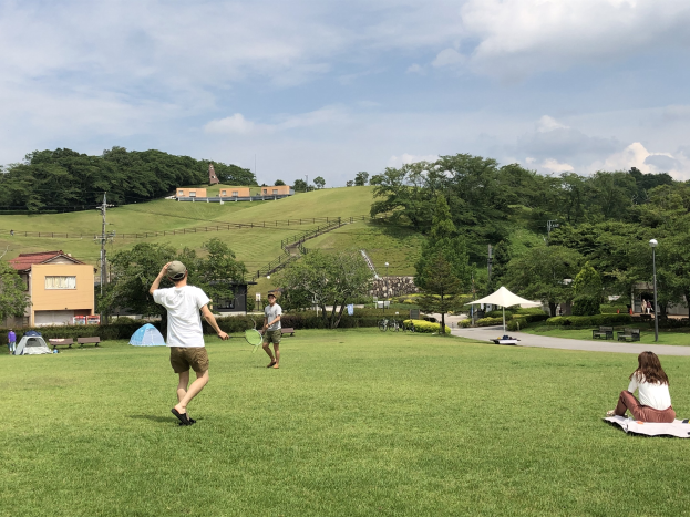 Gruppe von Menschen, die Badminton in einem Park spielt, mit Zelten, Laternenmasten und Gebäuden im Hintergrund.