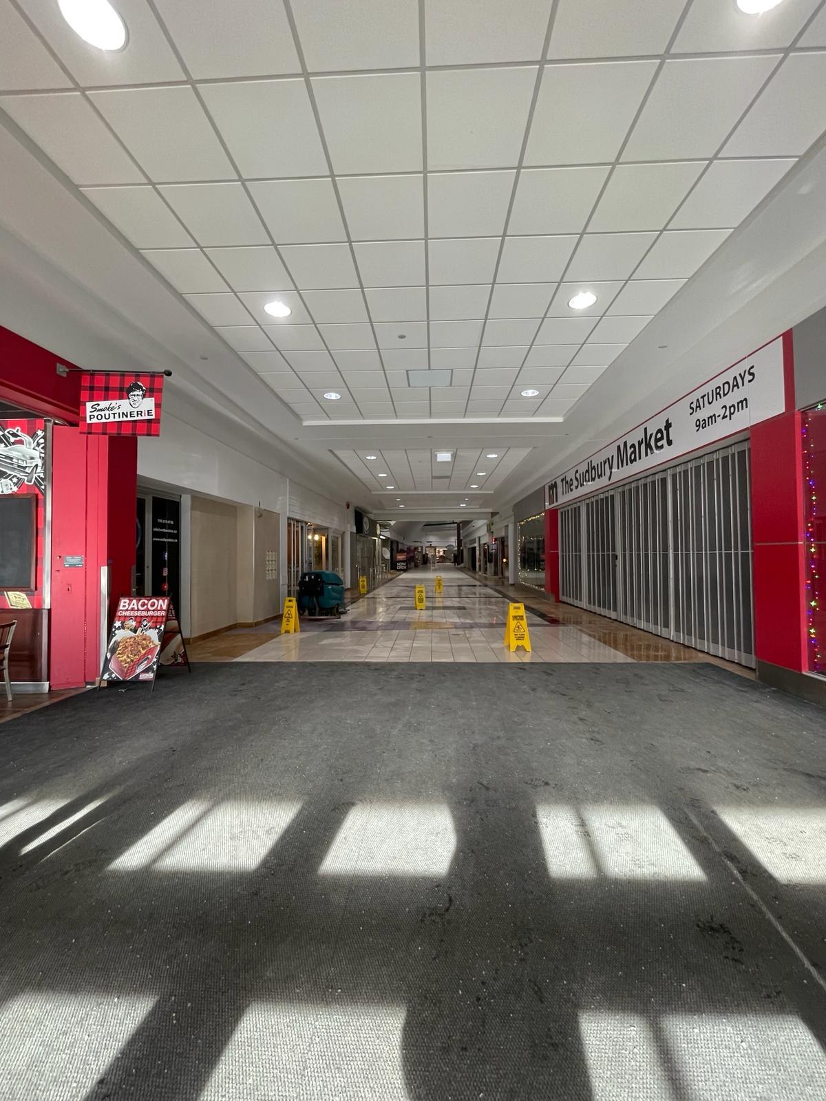 Innenseite eines Einkaufszentrums mit Deckenbeleuchtung, einem Stuhl und einer Tafel, auf der links "Sydbury Market - Vorher und Nachher" steht, sowie verstreuten Gegenständen rechts und auf dem Boden.