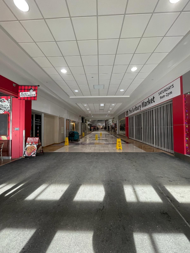 Innenseite eines Einkaufszentrums mit Deckenbeleuchtung, einem Stuhl und einer Tafel, auf der links "Sydbury Market - Vorher und Nachher" steht, sowie verstreuten Gegenständen rechts und auf dem Boden.