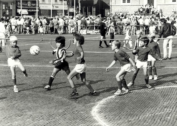 Schwarzes Bild von jungen Jungs, die auf der Straße Fußball spielen, umgeben von einer Zuschauermenge hinter einem Geländer und Gebäuden im Hintergrund.
