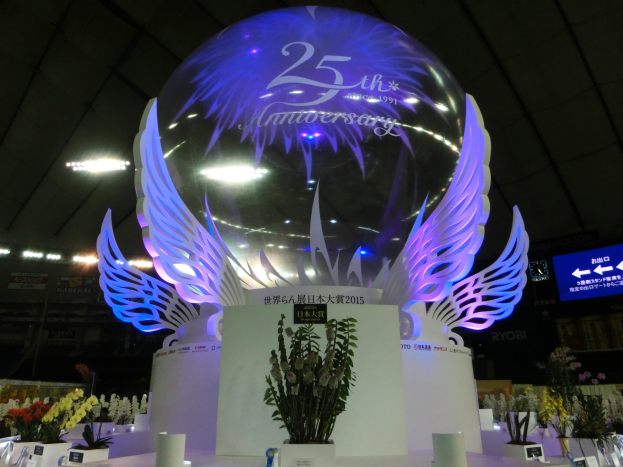 Ein großer Kristallball mit Flügeln oben drauf, umgeben von Blumenbouquets, Namensschildern und anderen Gegenständen auf dem Boden, vor einem festlichen Hintergrund mit Schildern, Lichtern und einem Bildschirm für eine 25-jährige Jubiläumsfeier.