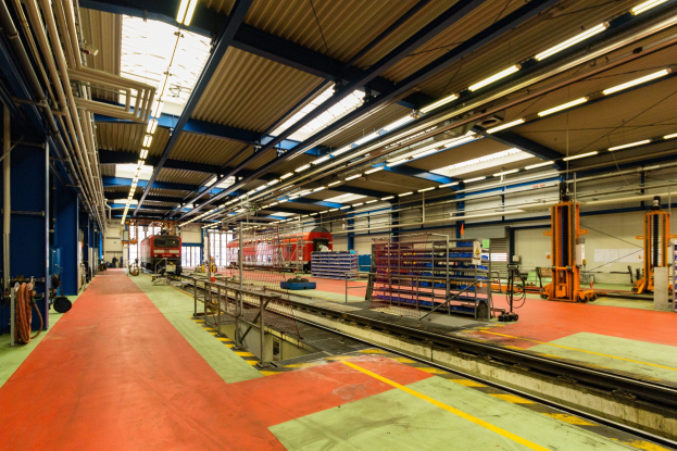 Großer Fabrikinnenraum mit rotem Teppich, Eisenbahnschienen, Geländern, Maschinen, Masten und Deckenleuchten, mit einem Zug im Hintergrund.