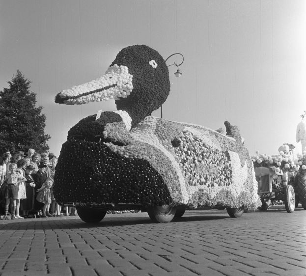 Schwarzes und weißes Foto eines Enten-Umzugswagens, der mit Blumen geschmückt ist und von Menschen umgeben ist