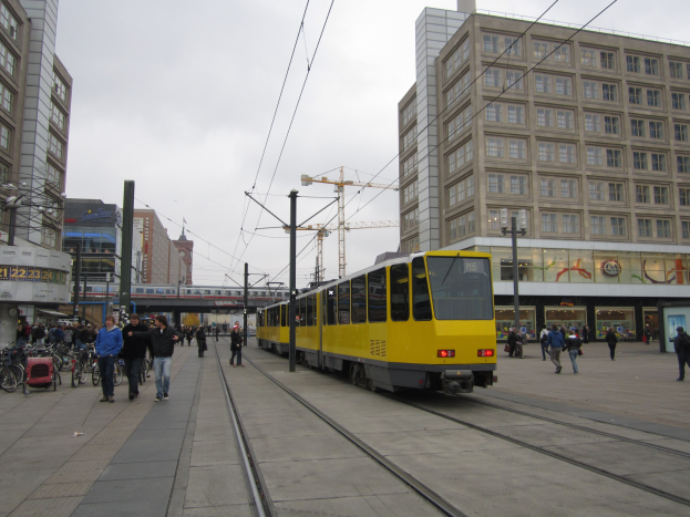 Gelber Straßenbahnwagen auf einer Stadtstraße mit Fußgängern, parkenden Fahrrädern auf dem Gehweg, Gebäuden, Strommasten, Oberleitungen, einem Baukran und Himmel im Hintergrund.
