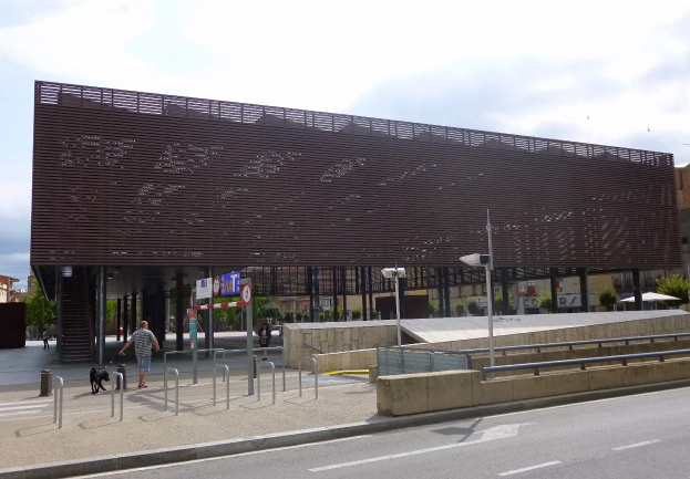 Großes modernes Gebäude mit zahlreichen Fenstern als neuer Eingang zur Stadtmitte, umgeben von Straßeninfrastruktur, Fahrzeugen, Fußgängern und Bäumen unter einem bewölkten Himmel.