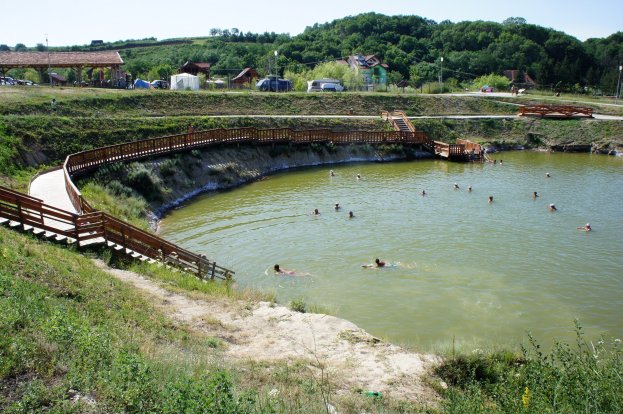 Gruppe von Menschen, die in einem Gewässer schwimmen, mit üppiger grüner Umgebung, einer Brücke mit Stufen, die zum Wasser führen, Schuppen, Fahrzeugen, Pfählen und einem klaren blauen Himmel im Hintergrund.