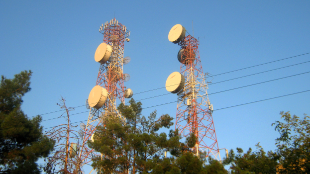 Zwei Mobilfunkmasten mit Antennen stehen zwischen Bäumen und Drähten vor einem klaren blauen Himmel.