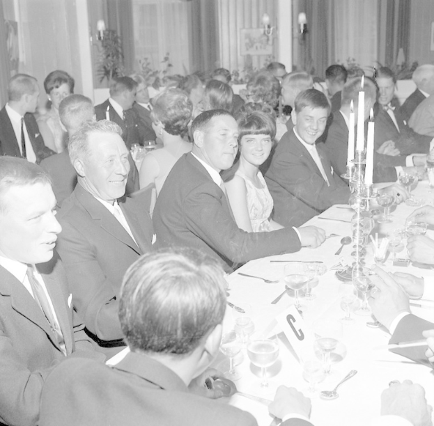 Eine Gruppe von Menschen sitzt um einen langen Tisch, lacht und genießt die Gesellschaft des anderen, mit Gläsern, Besteck, Kerzen und anderen Gegenständen auf dem Tisch und Vorhängen und Wandlampen im Hintergrund, die eine gemütliche Atmosphäre schaffen.