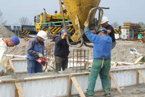 Männer mit Helmen bedienen einen Betonmischer auf einer Baustelle mit Fahrzeugen, Bäumen, Gebäuden und einem klaren blauen Himmel im Hintergrund.