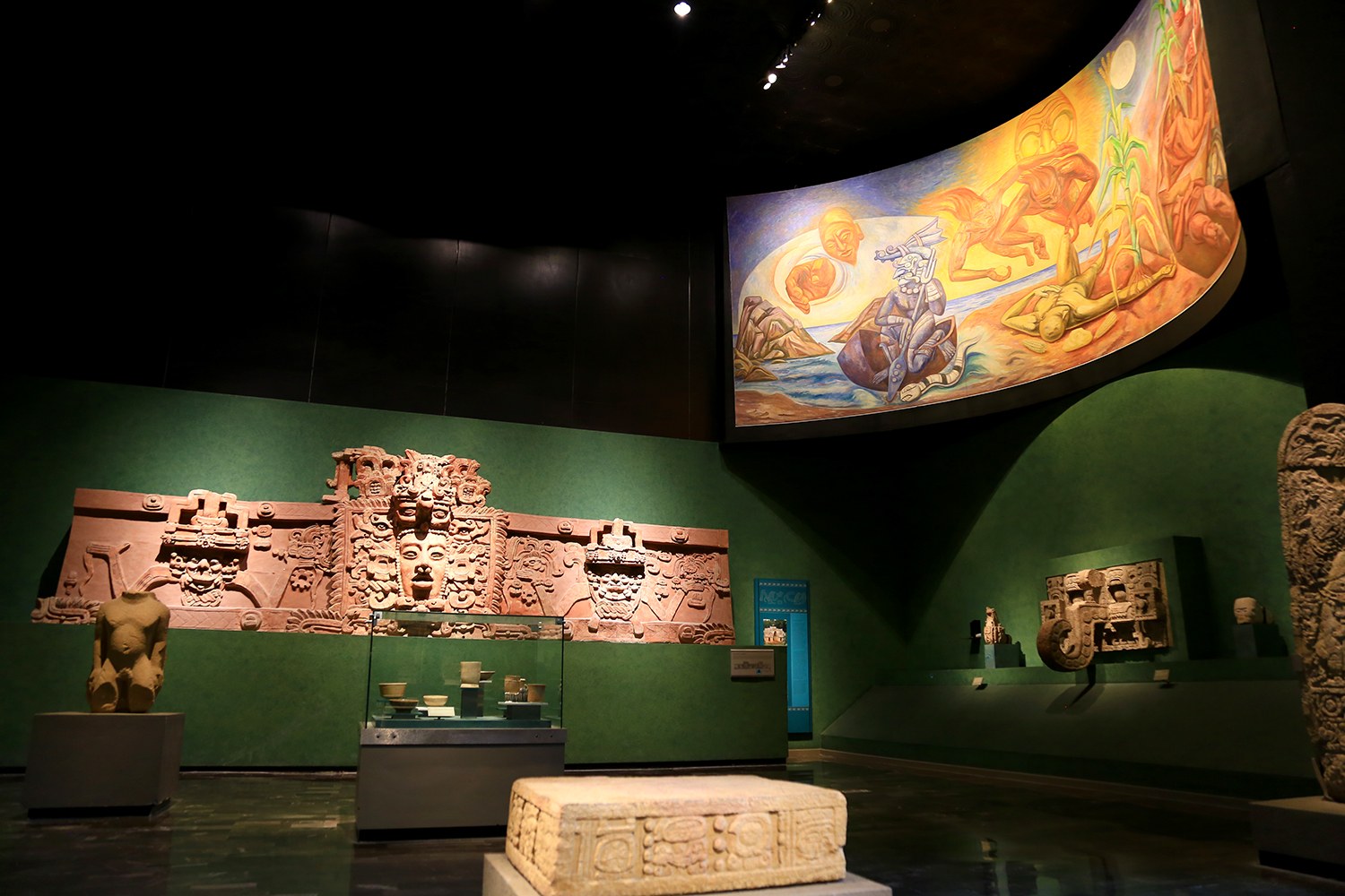Ausstellungsraum im Anthropologischen Museum von Mexiko-Stadt mit verschiedenen Kunstwerken, darunter Skulpturen, Tischgegenstände, eine Texttafel, eine Wandmalerei und Deckenbeleuchtung.