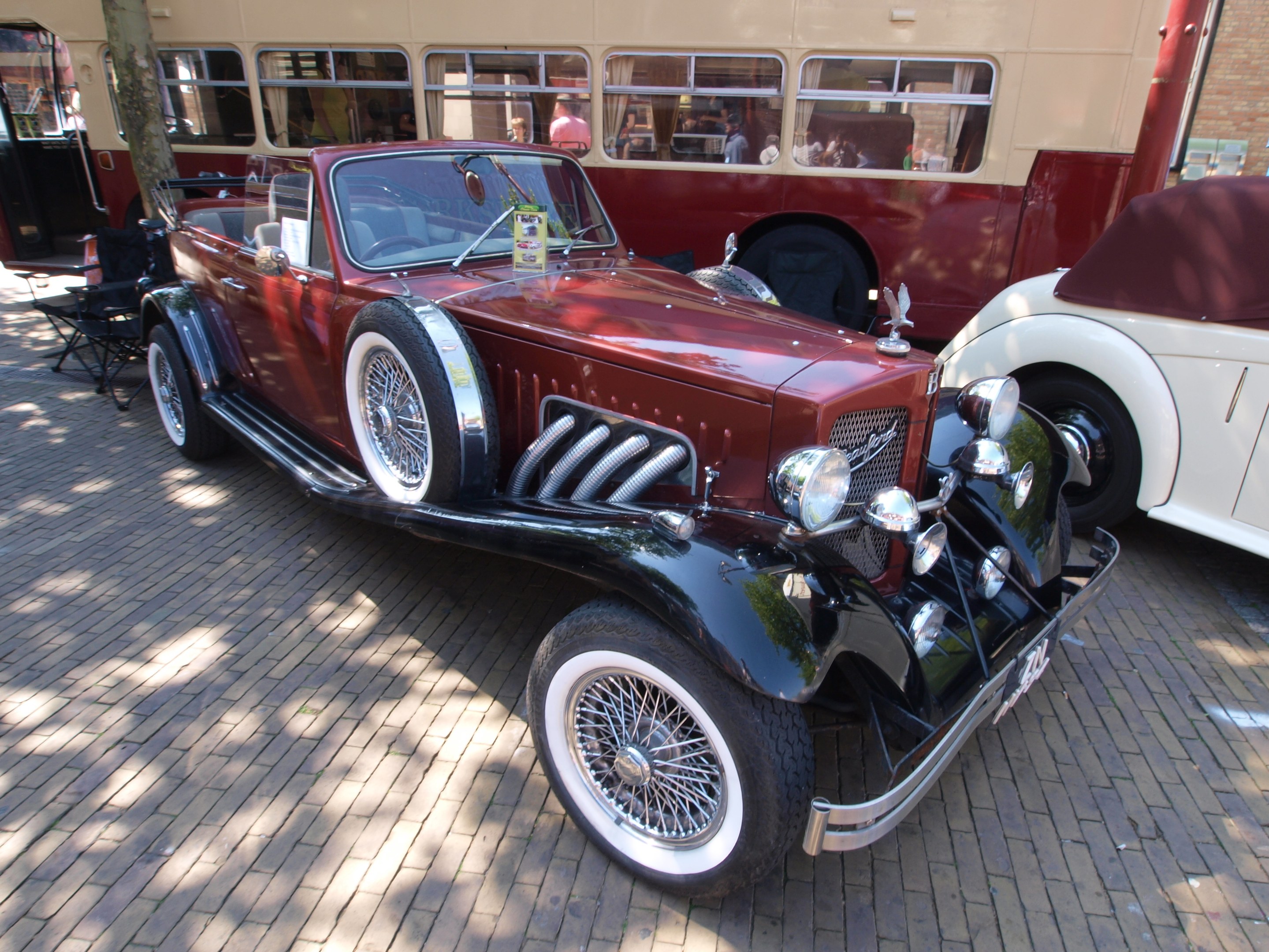Vintage-Auto neben einem Doppeldeckerbus auf einer Kopfsteinpflasterstraße geparkt, mit Stühlen und einem Baum im Vordergrund, einem Gebäude mit Fenstern im Hintergrund und Menschen im Bus sichtbar.